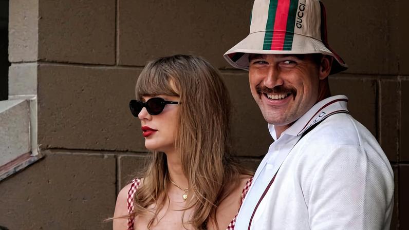 Mulher com vestido xadrez vermelho e branco e óculos escuros ao lado de homem sorridente usando chapéu bucket com listras verde e vermelha e camisa branca, encostados em parede bege.