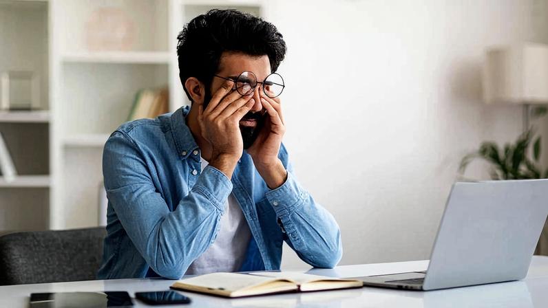 Homem sentado à mesa com laptop aberto, segurando o rosto com as mãos enquanto fala ao telefone. Mesa com caderno aberto, celular e ambiente interno claro ao fundo.