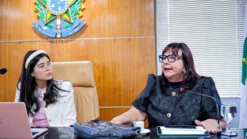 Duas mulheres sentadas em mesa de reunião com microfones. À esquerda, mulher jovem com cabelo longo, óculos e roupa clara observa a outra mulher, que fala e veste roupa escura. Ao fundo, brasão nacional brasileiro em parede de madeira e bandeira do Brasil à direita.