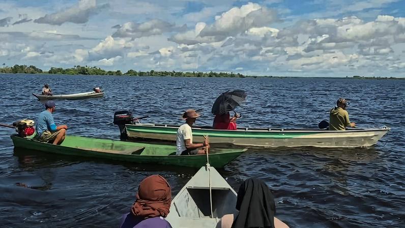 Cinco pessoas em três barcos pequenos no rio. Dois homens em barco verde à esquerda, uma pessoa com guarda-chuva em barco branco ao centro, e duas pessoas em barco ao fundo. Céu azul com nuvens dispersas.