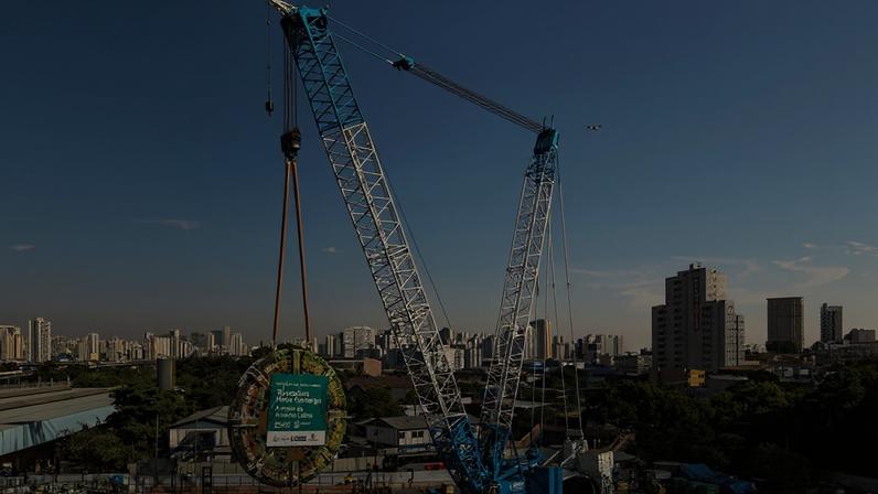 Guindaste azul e branco ergue componente circular grande em canteiro de obras na cidade. Edifícios altos e céu claro ao fundo.