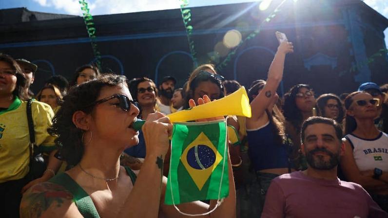 Grupo de pessoas vestindo camisetas da seleção brasileira em evento ao ar livre durante o dia. Mulher na frente usa óculos escuros e toca uma corneta amarela com uma pequena bandeira do Brasil pendurada. Outras pessoas ao fundo também vestem verde e amarelo, algumas com braços erguidos, sob céu azul com nuvens e decoração com bandeirinhas verdes e amarelas.