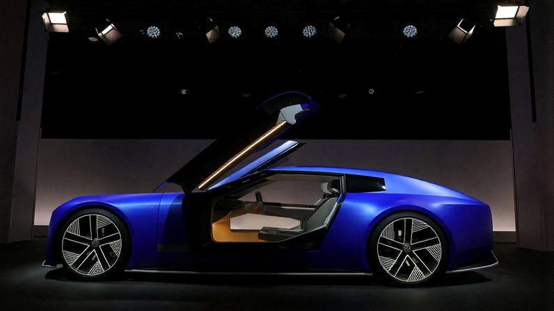 Carro esportivo azul metálico exibido em ambiente escuro com iluminação focada. Portas do tipo asa de gaivota estão abertas, revelando interior claro com assentos modernos e painel minimalista. Rodas têm design geométrico em preto e prata.