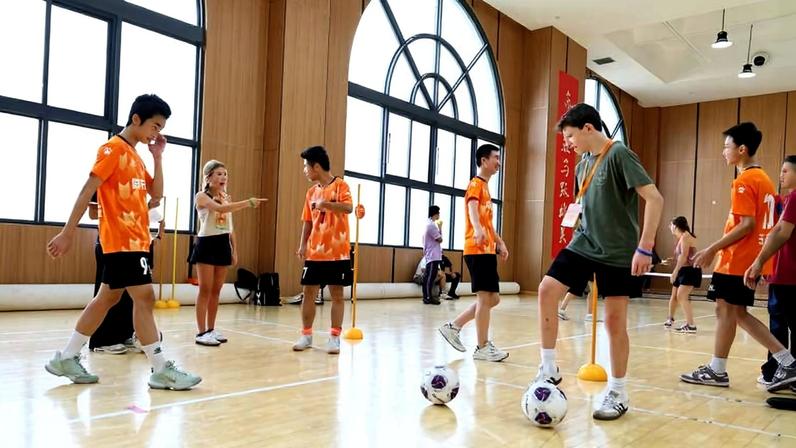 Seis jovens atletas com uniformes laranja e shorts pretos treinam em quadra coberta com piso de madeira. Dois deles controlam bolas de futebol próximas a cones laranja, enquanto uma mulher orienta e aponta para um dos jogadores. Grandes janelas arqueadas iluminam o ambiente interno.