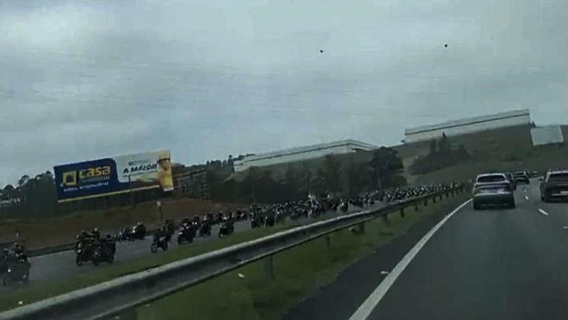 Longa fila de motociclistas parados no acostamento de uma rodovia de pista dupla em dia nublado. Veículos de passeio trafegam nas faixas principais à direita, enquanto a fila de motos se estende ao longo do gramado e da barreira de proteção à esquerda.