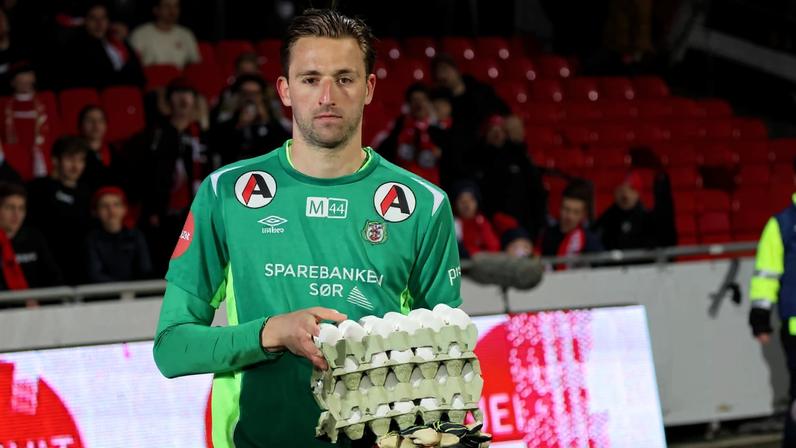 Goleiro vestido com uniforme verde segura três bandejas empilhadas de ovos durante partida de futebol. Torcida ao fundo em arquibancada vermelha.