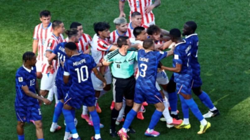 Jogadores de duas equipes de futebol, uma com uniformes listrados em vermelho e branco e outra com uniformes azuis, se enfrentam no campo. Árbitro tenta separar os atletas no centro da confusão, enquanto outros jogadores observam próximos.