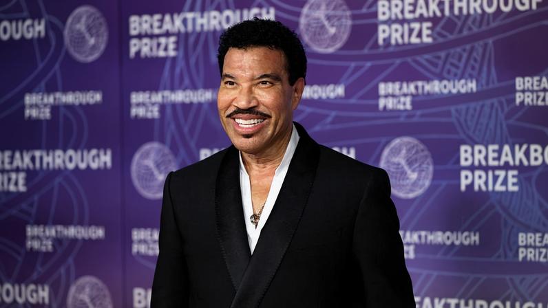 Homem de pele clara com cabelo preto encaracolado e bigode, veste terno preto com camisa branca, posando em frente a fundo roxo com texto 'Breakthrough Prize'.