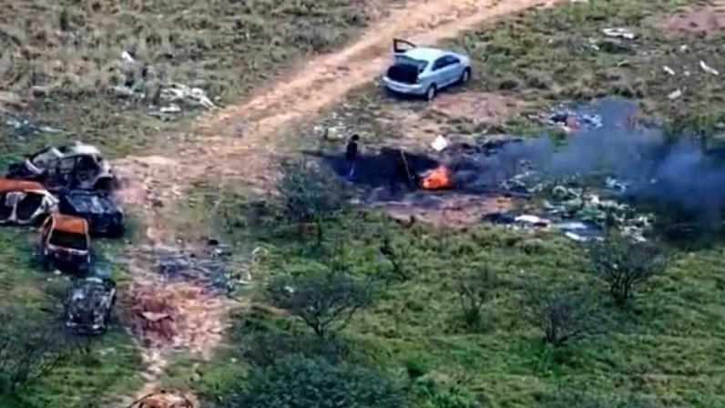 Área rural com quatro veículos queimados e destruídos à esquerda. Caminho de terra cruza a cena, com um carro branco estacionado à direita com porta traseira aberta. Pequena fogueira com fumaça preta visível próximo ao carro branco. Vegetação baixa e arbustos dispersos no entorno.
