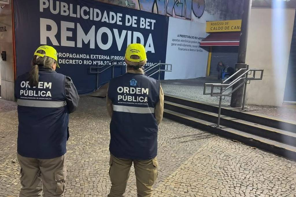 Agentes da prefeitura do Rio observando propaganda de bet tampada com aviso municipal