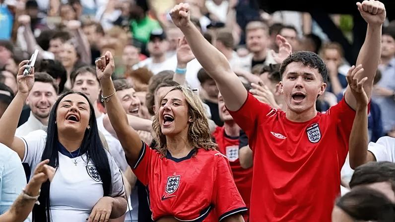 PA Media England fans celebrate in a pub garden. A crowd of men and women, in the foreground are two women and two men with their arms up wearing football t-shirts.