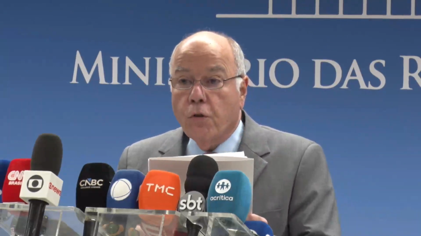 Mauro Vieira, de terno cinza e óculos, fala em coletiva de imprensa diante de microfones de veículos como CNN Brasil, GloboNews, CNBC, TMC, SBT e A Crítica, com o painel do Ministério das Relações Exteriores ao fundo.