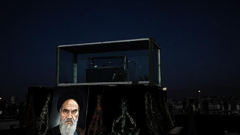 Um caixão verde dentro de uma estrutura de vidro sobre um palanque preto decorado com coroas de flores e um retrato do Aiatolá Khomeini. Pessoas em uniforme militar e um carro branco estão ao redor, com uma cidade ao fundo sob um céu azul