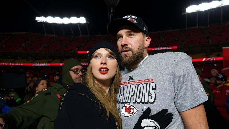 Taylor Swift, de touca preta e batom vermelho, abraça Travis Kelce, que usa boné e camiseta cinza com Champions escrito, em um estádio de futebol americano lotado e iluminado à noite