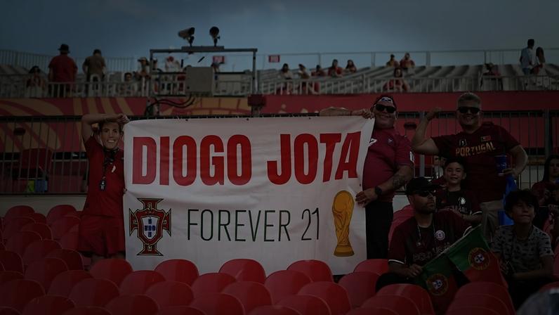 Torcedores portugueses em um estádio, segurando uma faixa branca com DIOGO JOTA FOREVER 21, o brasão de Portugal e um troféu dourado. Um homem e uma mulher sorriem, enquanto outros fãs, incluindo crianças, assistem. As cadeiras do estádio são vermelhas