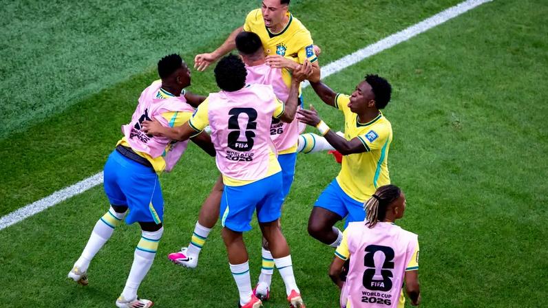 Jogadores de futebol brasileiros, vestindo camisas amarelas e coletes de treino rosa, se abraçam e comemoram em campo de grama verde, com uma linha branca visível