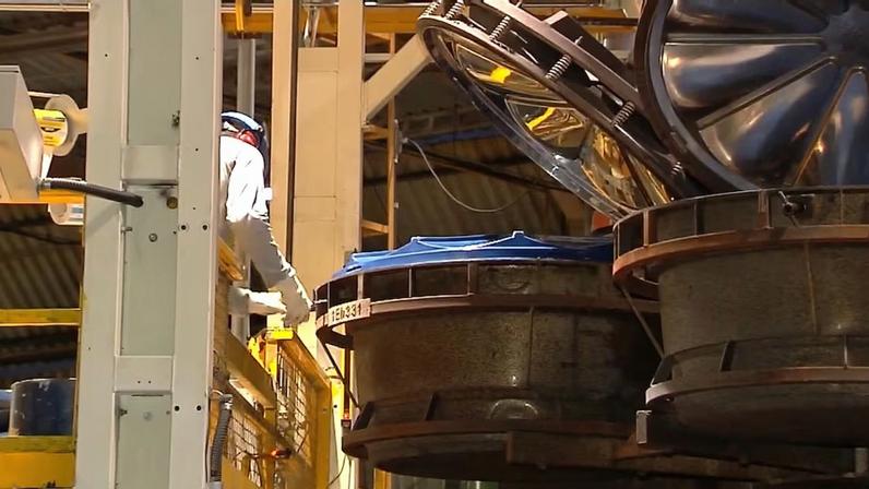 Vista de uma área industrial com grandes componentes metálicos circulares suspensos ou apoiados em uma linha de produção. Estruturas amarelas e colunas de suporte aparecem ao redor, sob iluminação interna do galpão.