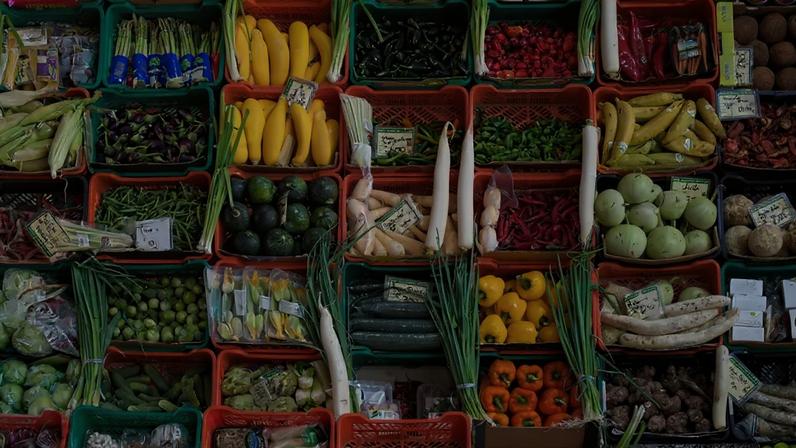 Exposição vibrante de frutas e vegetais frescos em caixas de plástico coloridas, incluindo milho, berinjelas, bananas, pimentões, abobrinhas, rabanetes, cebolinhas, cocos e aspargos, com etiquetas de preço visíveis.