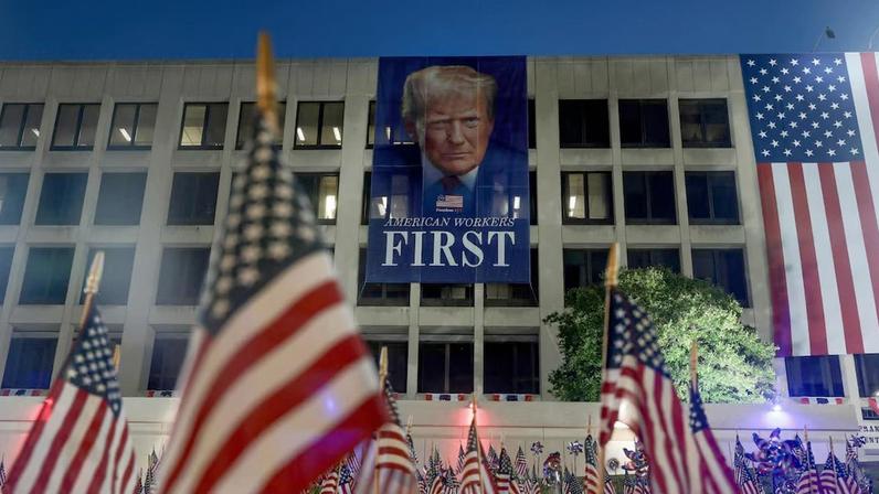 Bandeiras dos EUA estão fincadas no chão diante de um banner do presidente Donald Trump, suspenso no edifício do Departamento do Trabalho dos EUA, antes das festividades de 4 de julho