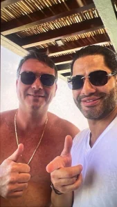 Foto mostra dois homens usando óculos escuros e fazendo sinal de positivo com as mãos. O homem à esquerda está sem camisa, usa corrente dourada e tem cabelo curto castanho. O homem à direita veste camiseta branca, tem barba e cabelo penteado para trás. Ao fundo, teto com estrutura de madeira e bambu.