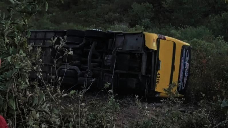Ônibus escolar amarelo tombado de lado em uma área de vegetação rasteira, com o chassi e as rodas expostos, após um acidente