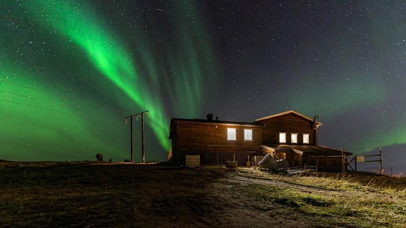 Foto colorida de aurora boreal de cor verde, captada em campo aberta com casa de campo em destaque - Metrópoles.