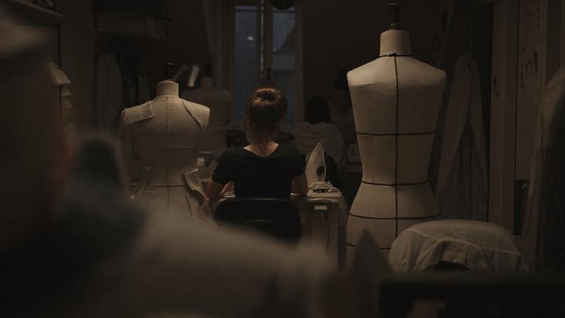 Pessoas trabalham em ateliê de moda. Mulher de costas passa roupa em mesa, manequins expostos ao redor.