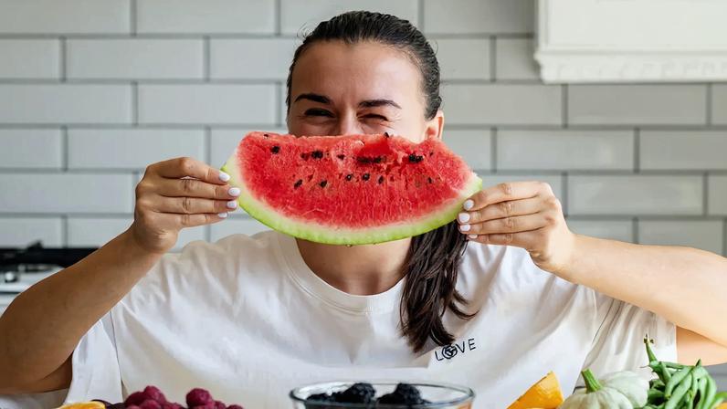 Foto colorida de mulher segurando uma fatia de melancia perdo do rosto, em frente a uma mesa cheia de frutas - Metrópoles.