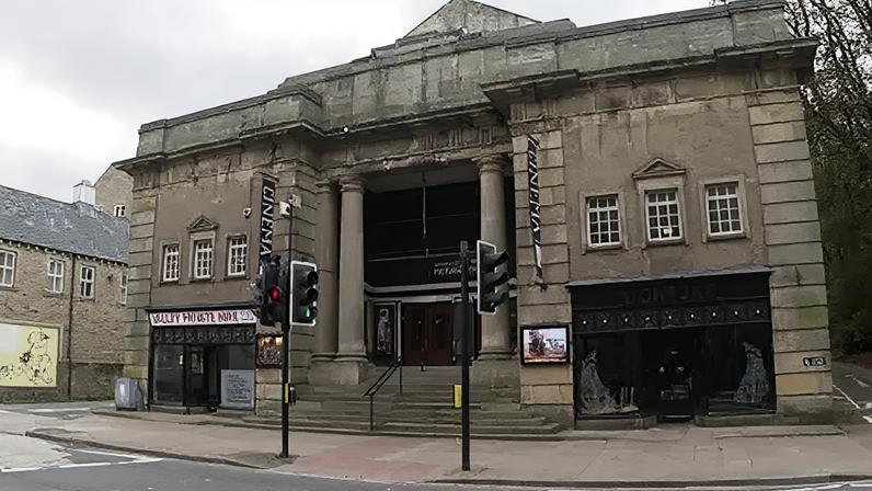 BBC An imposing grey stone building with two tall columns either side of the entrance doors in the middle. Both sides of the building's front have three sets of windows above separate shop fronts. The building sits next to a main road in front of a pedestrian crossing.