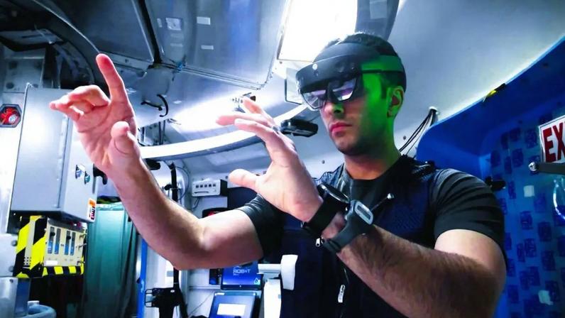 Um homem com óculos de realidade aumentada e camisa preta interage com uma projeção holográfica de um copo flutuante, dentro de um ambiente que lembra uma nave espacial, com painéis e adesivos no teto