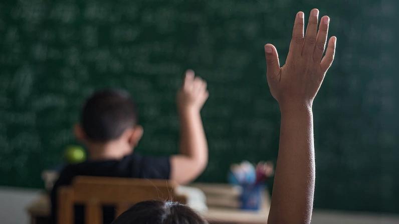 Foto colorida com zoom e falta de foco de mão infantil levantada em sala de aula - Metrópoles.