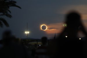 Coroa solar brilhante ao redor da Lua no céu escurecido ao entardecer, com nuvens alaranjadas ao fundo, silhuetas de pessoas, poste de luz e folhas de palmeira em primeiro plano