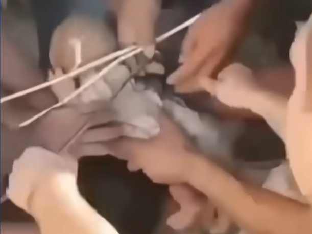 Diversas mãos retiram destroços para resgatar um bebê preso sob escombros, com fios e entulho visíveis ao redor.