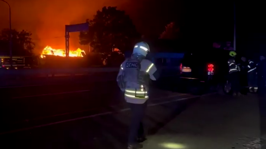 Bombeiro de costas observa grande incêndio ao longe, à noite, com viaturas e outros socorristas próximos ao local.