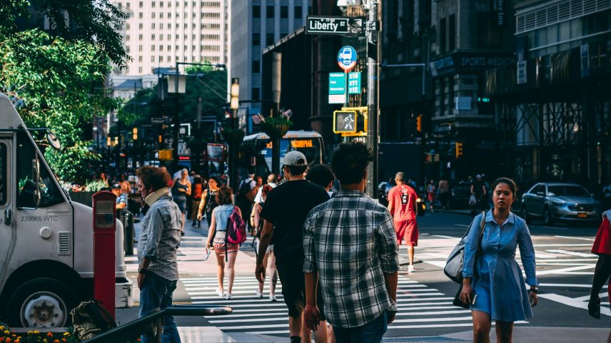 Rua urbana movimentada com pedestres caminhando na calçada, um jovem andando de skate, prédios altos ao fundo, faixa de pedestres e placa de rua indicando "Liberty St".