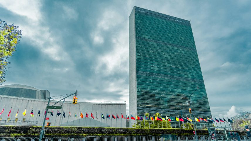 Fachada de um arranha-céu retangular revestido em vidro verde-azulado, com bandeiras de diferentes países hasteadas em fileira na base do prédio, sob céu parcialmente nublado.
