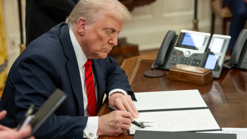 Donald Trump sentado à mesa do Salão Oval, curvado sobre um documento aberto, assinando com uma caneta preta. Ele veste terno azul-marinho, camisa branca e gravata vermelha com listras diagonais. Ao fundo, aparelhos telefônicos sobre a mesa e parte de outras pessoas em pé.