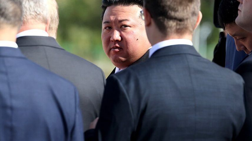 Kim Jong Un, de terno escuro, observado por seguranças e assessores ao seu redor durante evento externo.