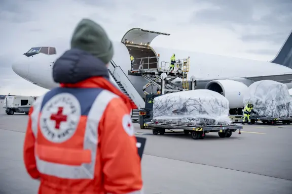 Funcionário de colete refletivo em cima de escada de embarque de avião de carga, com paletes cobertos por lona sendo transportados no pátio, e pessoa com uniforme da Cruz Vermelha Alemã em primeiro plano, de costas.