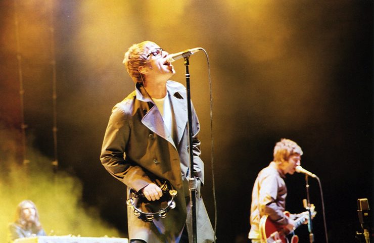 Liam Gallagher canta em um microfone usando um sobretudo cinza e óculos escuros, segurando um pandeiro, enquanto Noel Gallagher toca guitarra ao fundo, em um show do Oasis.