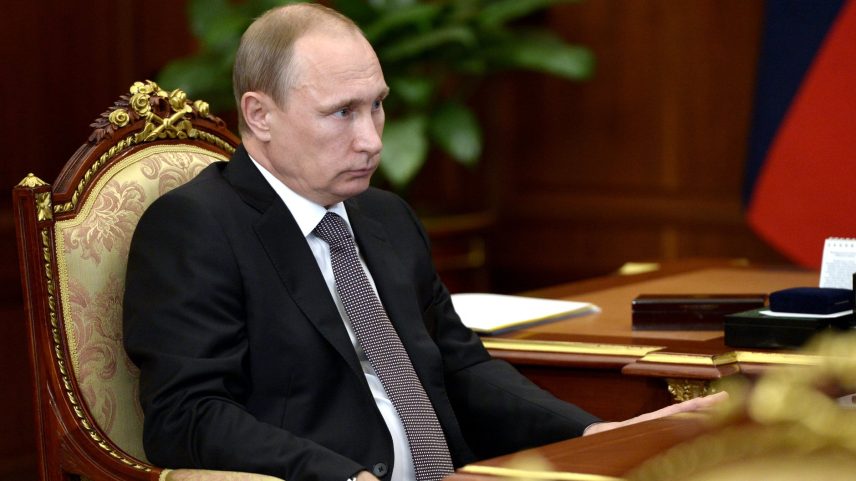 Vladimir Putin, de terno escuro e gravata, sentado em cadeira ornamentada durante reunião, com bandeira da Rússia ao fundo