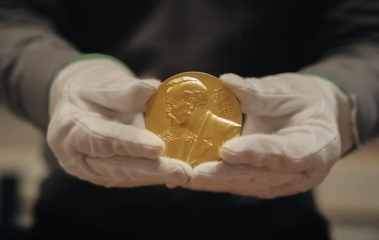 Mãos com luvas brancas seguram uma medalha dourada com o perfil em relevo de um homem de barba e a inscrição "Alfr. Nobel", diante de um fundo escuro e desfocado.