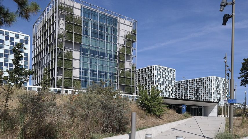 Conjunto de prédios modernos sob céu azul. À esquerda, um edifício envidraçado tem a fachada coberta por uma estrutura metálica com vegetação. Ao fundo, aparecem dois blocos brancos com janelas quadradas em padrão irregular. Em primeiro plano, uma calçada cercada por mato seco leva até a entrada.