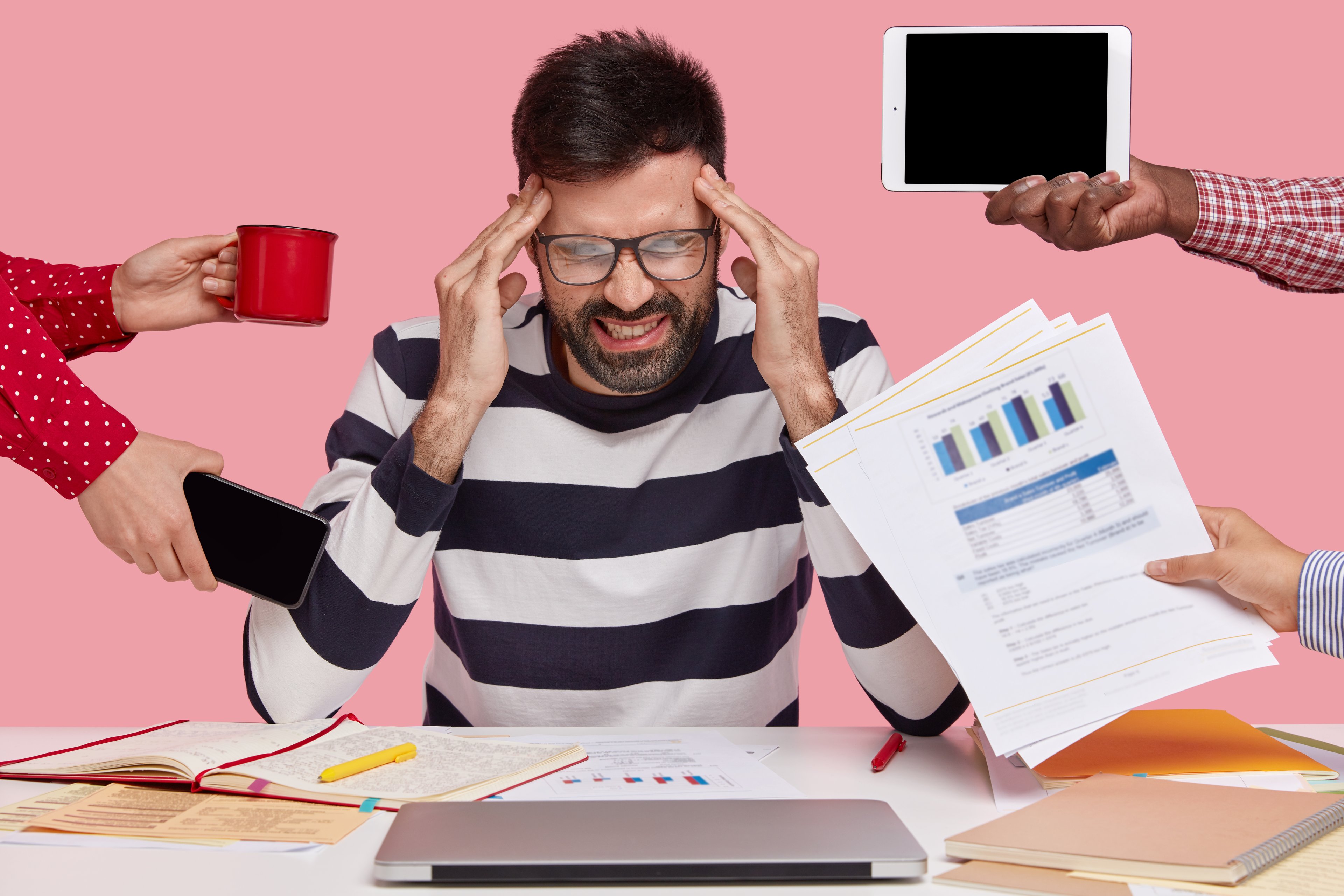 Marcas enfrentam dilema entre crescimento e valores essenciais para o consumidor Homem de marketing sobrecarregado esfrega as têmporas, precisa se concentrar e trabalhar no relatório financeiro, focado para baixo, segura uma caneca de café quente, papéis com gráficos, touchpad e celular, posa em ambiente interno. (Foto: Freepik)