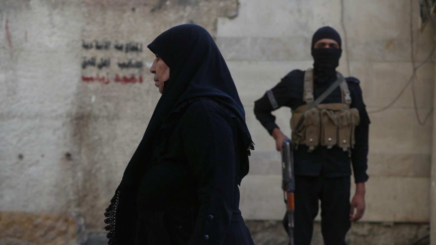 Uma mulher síria passa por um homem da segurança do governo sírio, do lado de fora do santuário de al-Sayydah Zeinab, neta do Profeta Maomé, onde fiéis xiitas participam do ritual de Ashoura, ao sul de Damasco, Síria, quinta-feira, 3 de julho de 2025. (Foto: AP Photo/Omar Sanadiki)