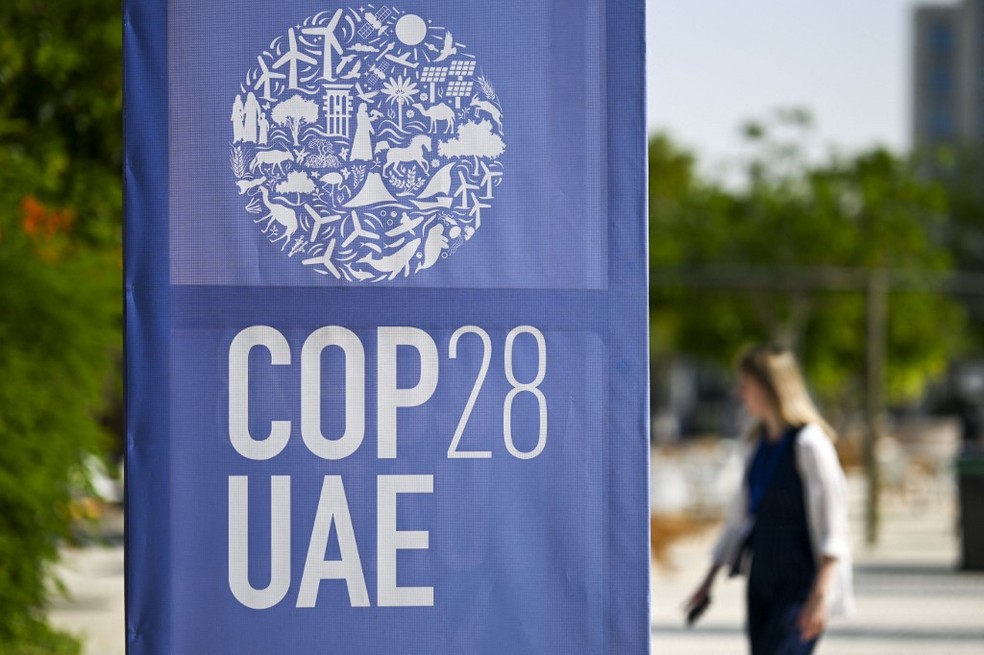 COP é essencial para o futuro do clima e das políticas ambientais globais Uma mulher caminha perto de um painel com o logo da COP 28 exibido em Dubai. (Foto: Jewel SAMAD/AFP)