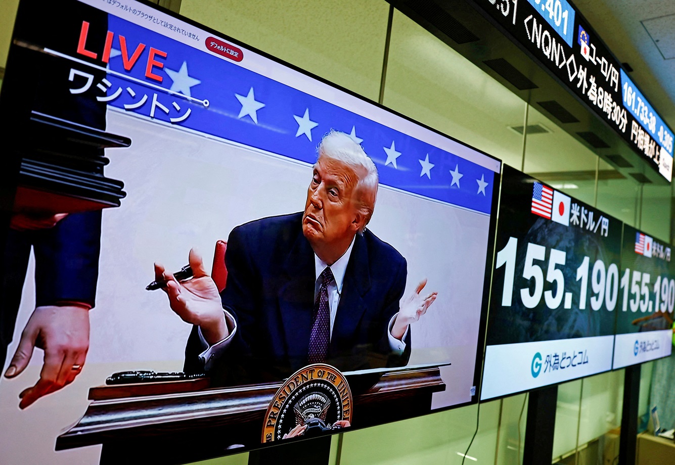 Uma tela transmitindo imagens ao vivo mostra o presidente dos EUA, Donald Trump, gesticulando enquanto assina ordens executivas ao lado de telas que exibem a taxa de câmbio atual do iene japonês em relação ao dólar americano na sala de operações da empresa de câmbio Gaitame.com, em Tóquio, Japão, 21 de janeiro de 2025. (Foto: REUTERS/Issei Kato/FOTO DE ARQUIVO)