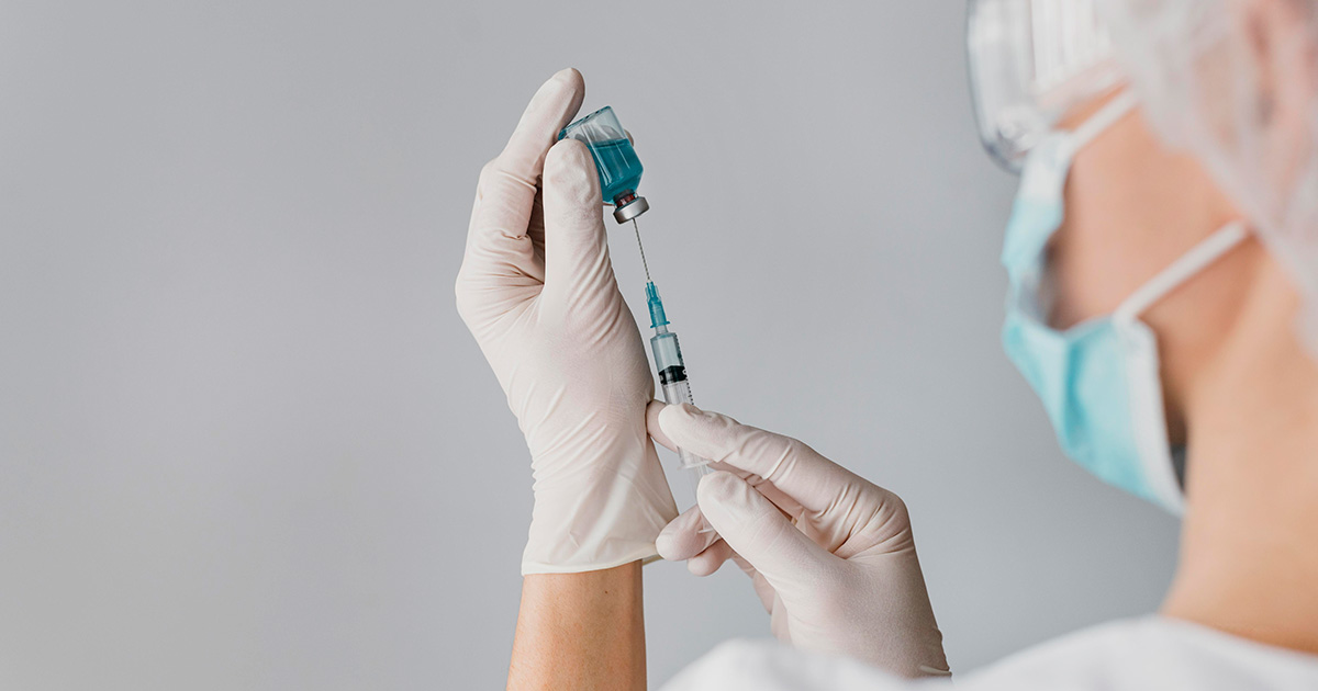 Vacina contra covid da USP avança com resultados promissores em nova fase de testes Pessoa de perfil, com touca e máscara cirúrgica, segura um frasco de conteúdo azul e insere uma seringa nele. (Foto: USP Imagens)