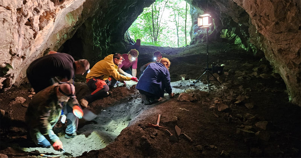 Seis pessoas agachadas trabalhando com ferramentas na escavação do solo no interior de uma caverna (Foto: USP Imagens)