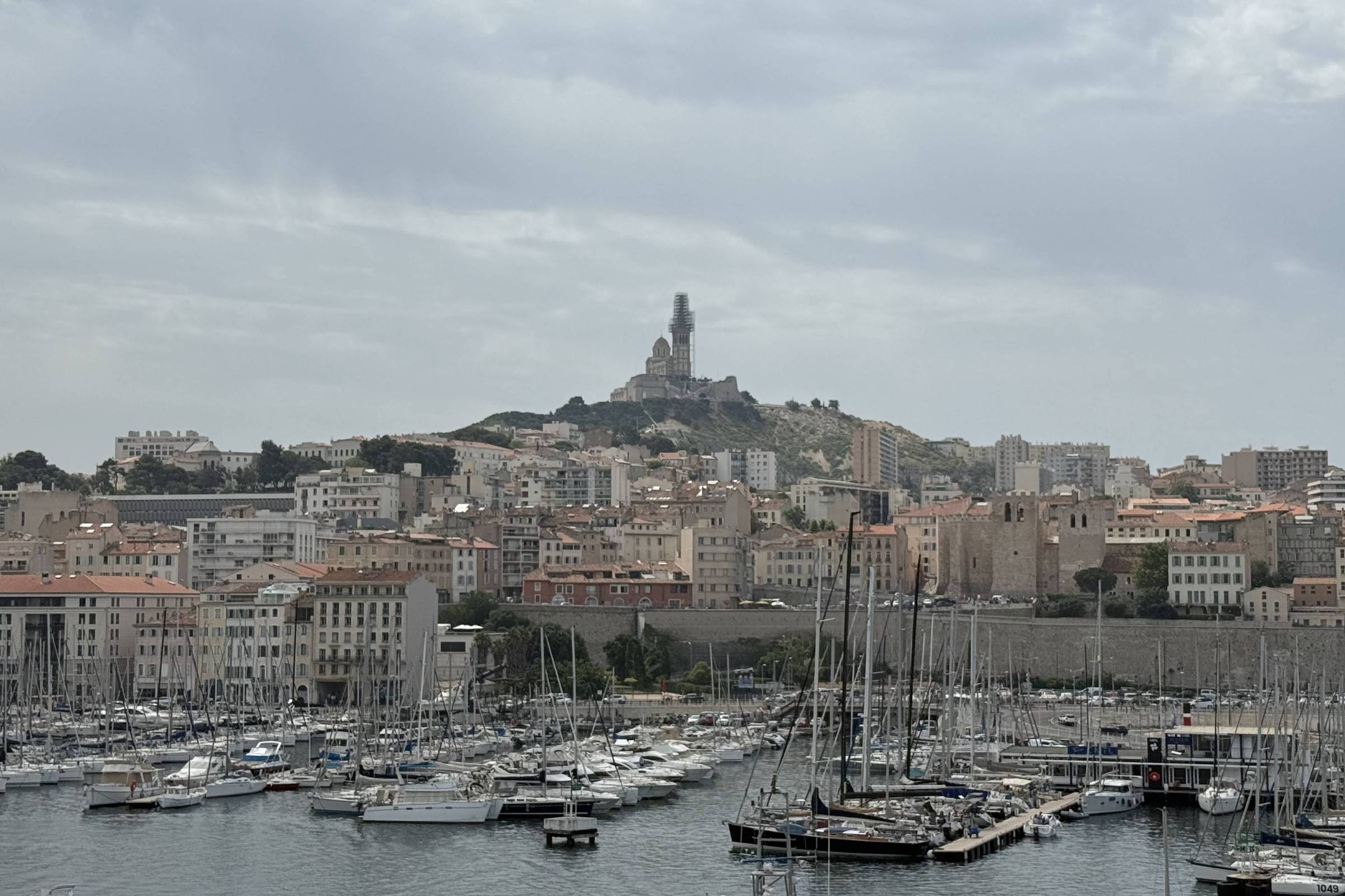 Marselha se destaca como opção acessível e vibrante em relação a Paris A cidade de Marselha, com o porto antigo, no primeiro plano, e a Basílica Notre Dame de la Garde, no fundo (Foto: Diogo Bercito/Folhapress)